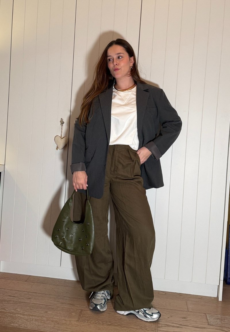 Femme portant un blazer noir, une chemise blanche, un pantalon large olive, des baskets argentées, tenant un sac à main vert olive, debout contre un mur blanc à panneaux.