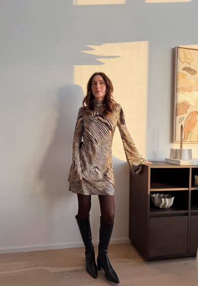 Mujer de pie en el interior, vistiendo un vestido de manga larga con estampado de cebra, medias negras y botas negras hasta la rodilla, apoyada en un armario de madera.