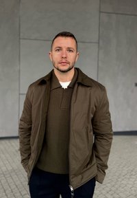 Mężczyzna z krótkimi włosami i brodą, ubrany w br�ązową kurtkę i sweter, stoi oparty o szarą, kafelkowaną ścianę, z rękami w kieszeniach.