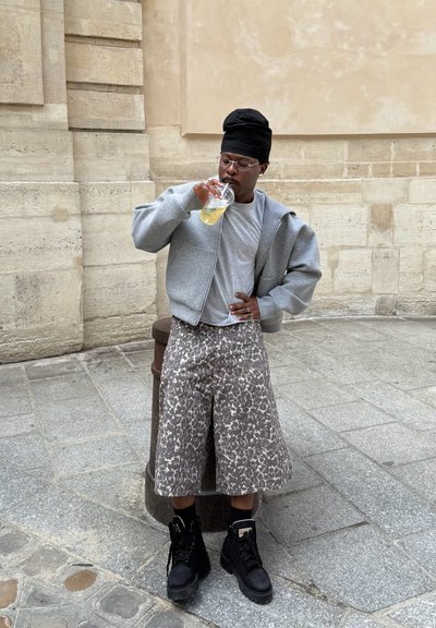 Sudadera gris con cremallera sobre una camiseta gris, pantalones cortos de diseño en marrón y beige, y botas negras con cordones. Bebiendo de un vaso transparente.