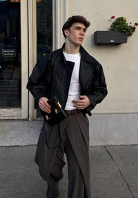 Veste en cuir noire par-dessus un t-shirt blanc, associée à un pantalon gris à jambes larges et un petit sac à main noir avec des accents dorés.