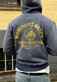 Felpa blu navy con grafica gialla con il testo "NEWPORT BEACH BOARD RENTAL" e palme. Maniche e orlo a coste, vestibilità rilassata, texture morbida.