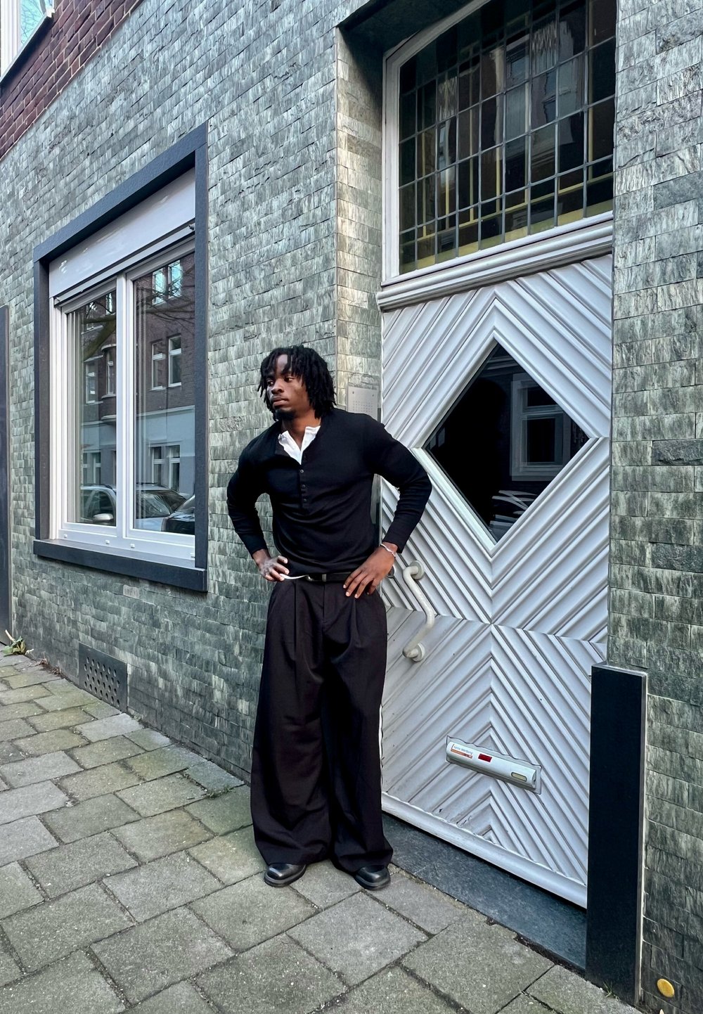 Homme aux dreadlocks portant des vêtements noirs et blancs, debout les mains sur les hanches à côté d'une porte moderne à motifs sur un trottoir en ville.