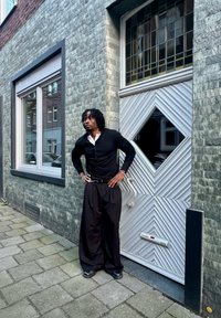 Homme aux dreadlocks portant des vêtements noirs et blancs, debout les mains sur les hanches à côté d'une porte moderne à motifs sur un trottoir en ville.