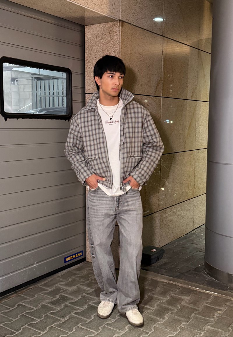 Jeune homme portant une veste à carreaux, une chemise blanche, un jean gris et des baskets blanches, debout près d'une porte de garage grise et d'un mur carrelé la nuit.