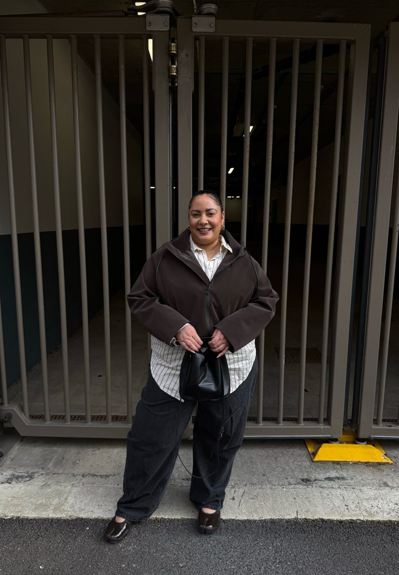 Femme souriante debout devant une grande porte métallique grise, portant une veste marron, une chemise rayée, un pantalon noir et tenant un sac noir.