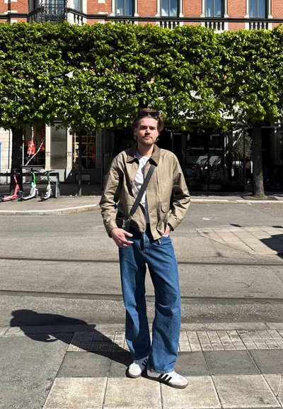 Chaqueta beige claro, camisa blanca, jeans azules y zapatillas blancas con rayas negras. De pie en una calle con vegetación y scooters de fondo.