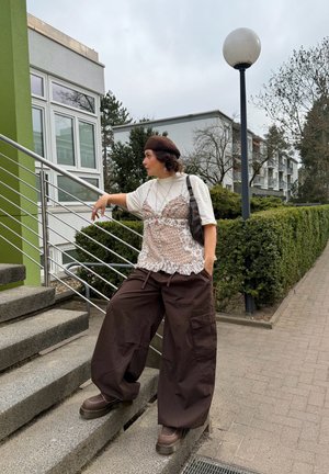 Persona con boina marrón, pantalones marrones extragrandes y camiseta beige con estampado apoyada en una barandilla metálica junto a unas escaleras exteriores cerca de un edificio verde.