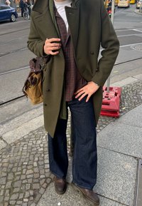 Casaco tipo peacoat verde-olive, camisa xadrez castanha, jeans escuros de perna larga, botas castanhas e uma bolsa castanha de ombro. Fundo de calçada de pedras.