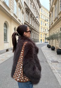 Veste en fausse fourrure marron avec une apparence texturée, associée à un sac en cuir marron orné de clous métalliques. Arrière-plan visible d'une rue de la ville.