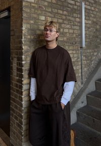 T-shirt marron surdimensionné à manches courtes superposé à une chemise blanche à manches longues, associé à un pantalon foncé ample et des chaussures décontractées.