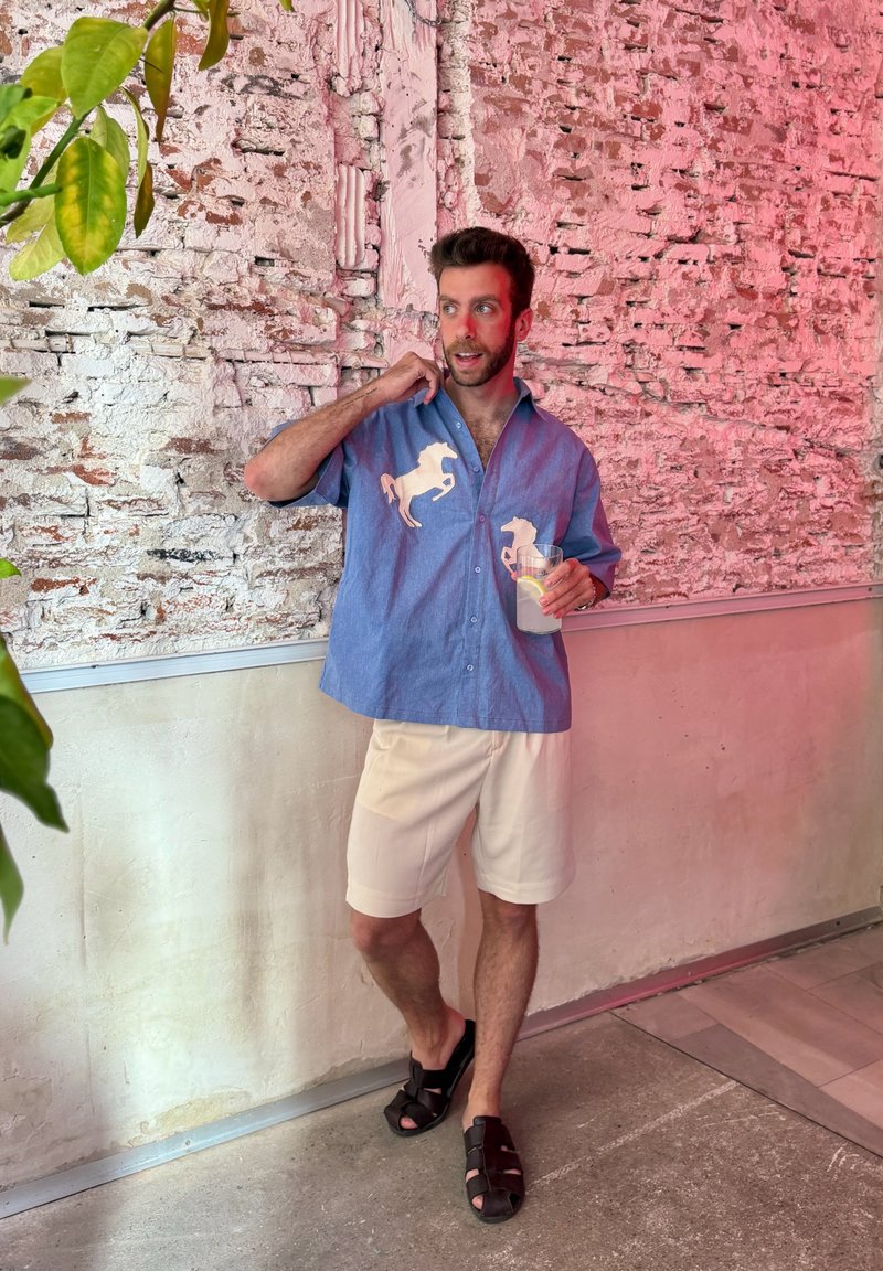 Homme en chemise bleue avec des motifs de chevaux, short beige et sandales noires, debout en train de boire de la limonade près d'un mur en briques blanc et rose.