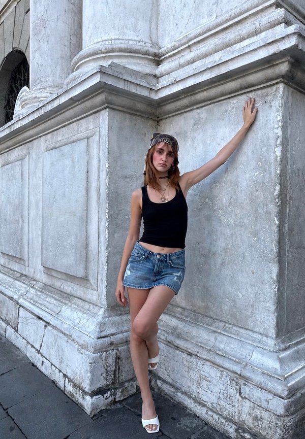 Ung kvinde iført sort tanktop, denimshorts og hvide hæle, der læner sig med en arm mod et tekstureret stenkrogen på en byens gade.