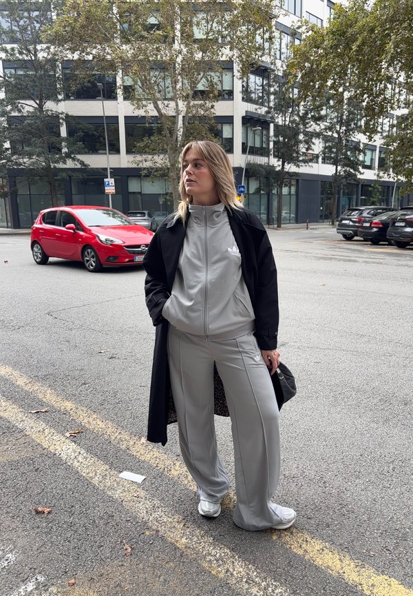 Light grey tracksuit with a zip-up jacket and matching trousers, black overcoat, and white sneakers. Urban setting with parked cars in the background.