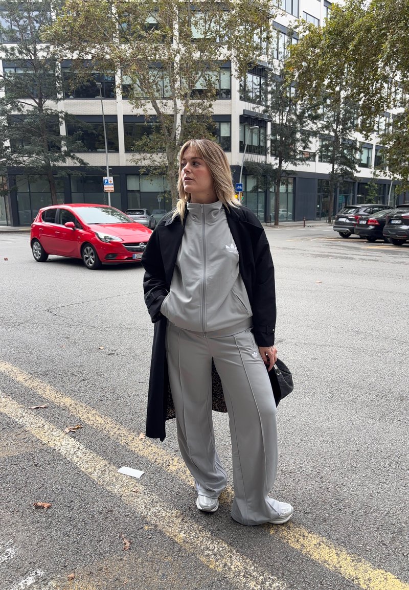Lys grå treningsdress med glidelåsjakke og matchende bukser, svart ytterjakke og hvite sneakers. Byrådmiljø med parkerte biler i bakgrunnen.