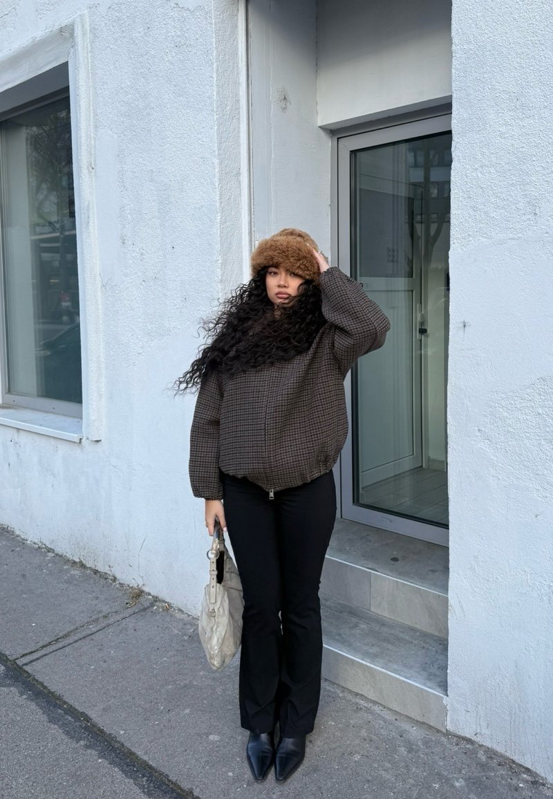 Γυναίκα με μακριά κατσαρά μαλλιά, φοράει καφέ αφράτο καπέλο, καρό σακάκι, μαύρο παντελόνι και μπότες, κρατώντας μια ανοιχτόχρωμη τσάντα δίπλα σε ένα λευκό κτίριο.