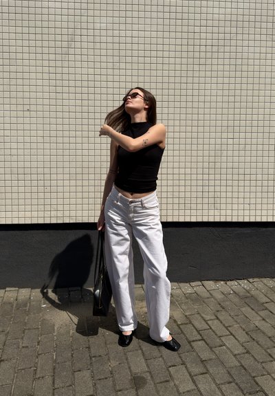 Mujer con el pelo largo y castaño que lleva gafas de sol, top negro sin mangas, pantalones blancos y zapatos negros, y está de pie en la acera junto a una pared alicatada.
