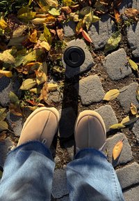 Pés calçando chinelos castanhos e jeans azuis a pisar um pavimento de calçada com folhas de outono amarelas e castanhas espalhadas e uma tampa de copo de café preta.