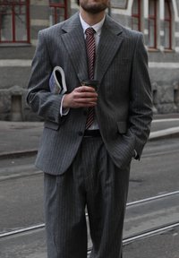 Grijs krijtstreeppak met een gestructureerde blazer en losvallende broek. Houdt een koffiekopje vast. Witte overhemd en rood gestreepte stropdas zichtbaar.