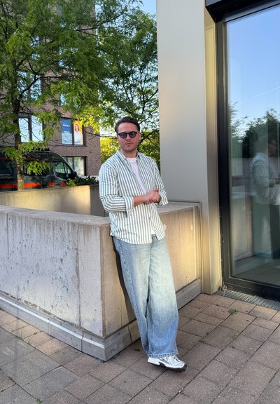 Camisa de botón a rayas en verde y blanco, vaqueros holgados azul claro y zapatillas blancas. De pie contra un pilar de concreto.