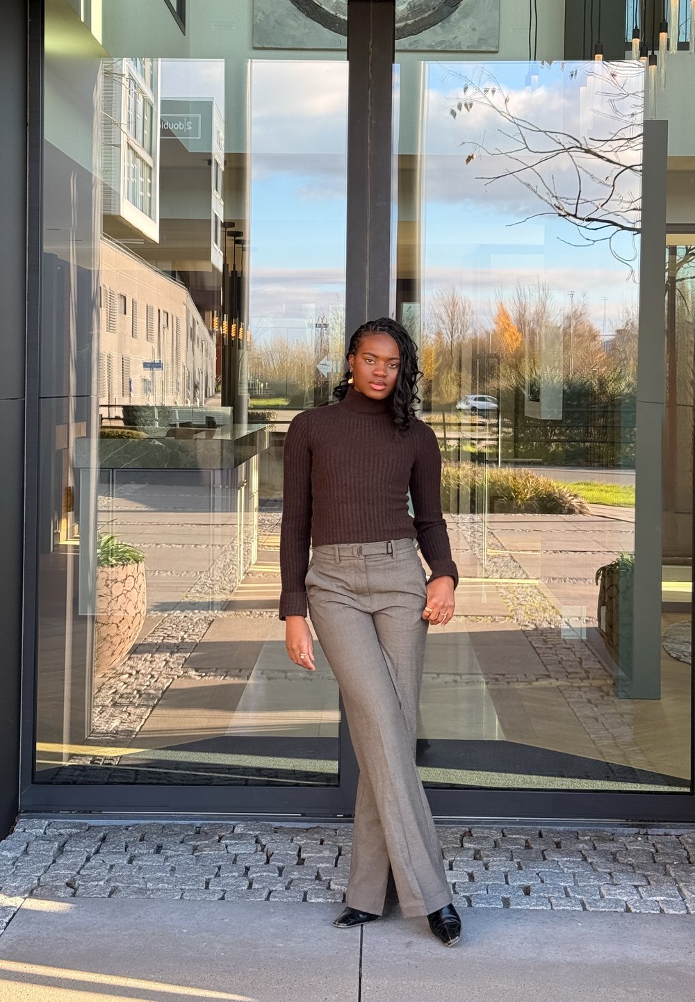 Brauner, gerippter Rollkragenpullover, graue, maßgeschneiderte Hosen und schwarze High Heels. Die Szenerie zeigt einen modernen Glaseingang und einen Boden mit Mustern aus Steinfliesen.