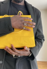 Osoba v černém kabátu a košili drží jasně žlutou nákupní tašku Swarovski, na rukou má stříbrné prsteny ve tvaru pavouka a geometrického designu.