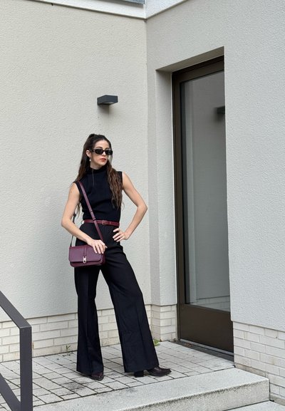 Mujer con top sin mangas negro y pantalones con cinturón y bolso burdeos, lleva gafas de sol, de pie cerca de la entrada de un edificio moderno.