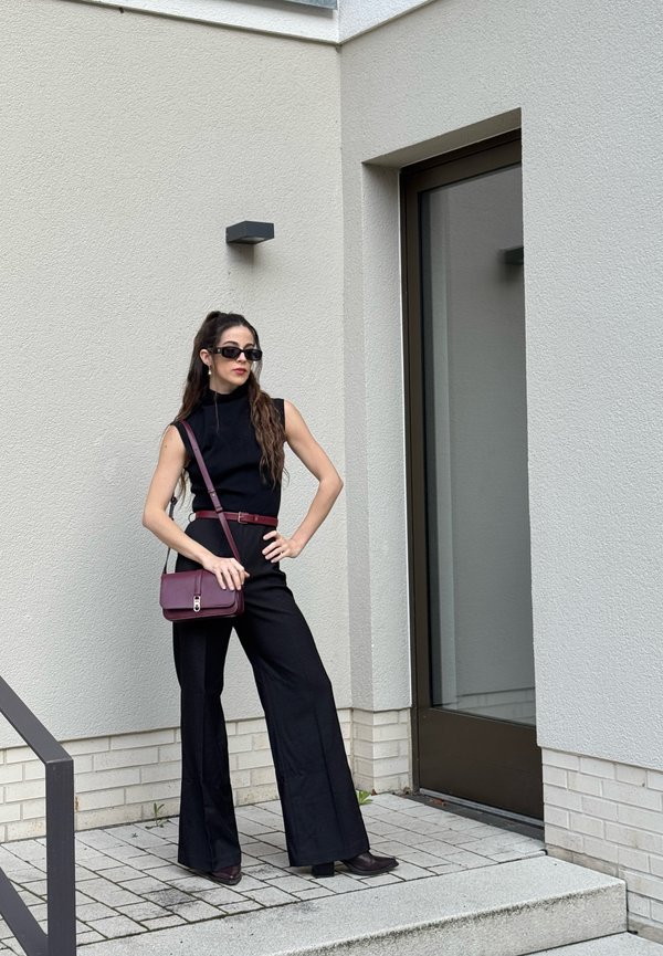 Frau in schwarzem ärmellosem Oberteil und Hose mit bordeauxrotem Gürtel und Tasche, trägt Sonnenbrille, steht in der Nähe eines modernen Gebäudeeingangs.