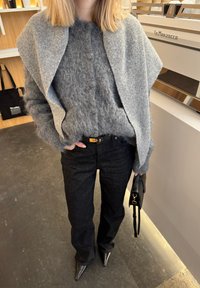 Personne portant un pull gris duveteux avec un châle assorti, un pantalon noir, des chaussures noires pointues, tenant un sac à main noir à l'intérieur près d'une machine à café.