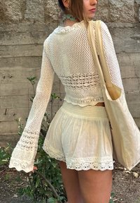 Bela kvačkana majica s dugimi rokavi in 
rokavi v zvončasti obliki ter valovitim robom, v kombinaciji s svetlo obarvanim plisiranim krilom z čipkastim robom. Bež torba.