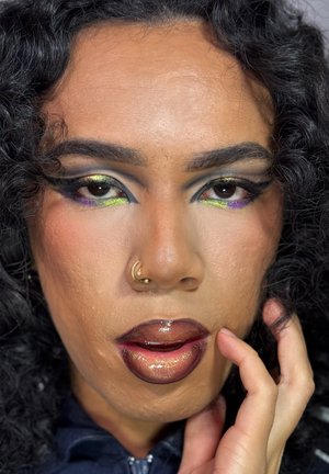 Primer plano de un rostro con cabello oscuro y rizado; maquillaje audaz con sombras de ojos verdes y púrpuras, labios marrones brillantes y un aro dorado en la nariz.
