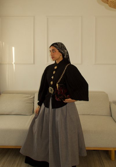 Blusa negra plisada con grandes botones dorados, combinada con una falda gris. Los accesorios incluyen un bolso floral y un pañuelo de cabeza estampado.