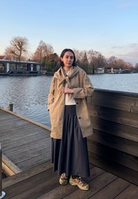 Μπεζ αδιάβροχο παλτό με κατακόρυφο γιακά, φορεμένο πάνω από ένα γκρι πουλόβερ και μαύρη μακριά φούστα. Καφέ και πράσινα αθλητικά παπούτσια με κορδόνια.