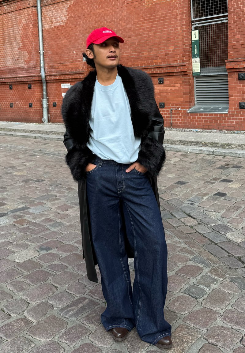 Black faux fur coat, light blue t-shirt, wide-leg dark denim jeans, and brown shoes, with a red cap in an outdoor urban setting.
