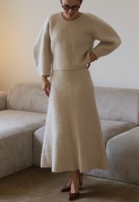 Bežani, baršunasti pleteni pulover s širokim rukavima u kombinaciji s odgovarajućom midi suknjom. Odjeća se odlikuje jednostavnom, ravnom siluetom i elegantnim potpeticama.
