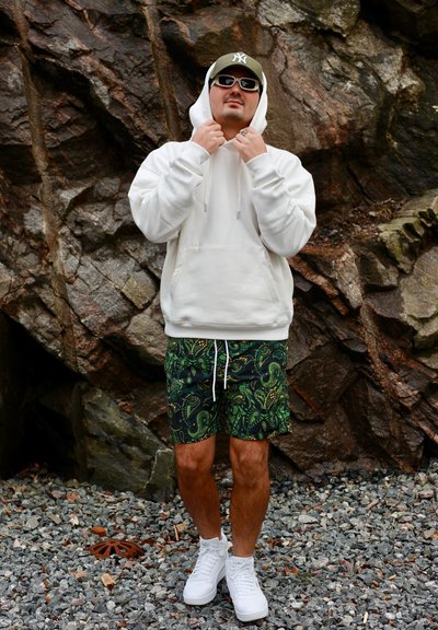 Hombre con sudadera blanca con capucha, pantalones cortos verdes con estampado, zapatillas blancas, gafas de sol y gorra oliva, de pie sobre terreno rocoso junto a un gran muro de piedra.