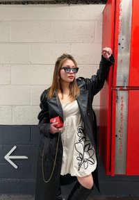 Femme en manteau en cuir noir, robe blanche à imprimé floral noir, lunettes de soleil et bottes montant jusqu'aux genoux s'appuie contre une armoire rouge d'extincteur.
