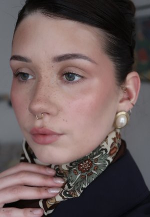 Mujer joven con pecas, que lleva un pañuelo con estampado, un aro dorado en el septum, un gran pendiente de perla y ropa azul marino, sosteniendo su pañuelo.