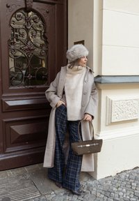 Manteau en laine beige, pull léger en maille, pantalon large à carreaux, chapeau en fausse fourrure gris et sac à main foncé avec accents dorés. Porte unie et mur texturé.