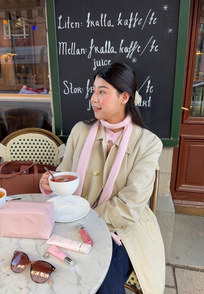 Mujer con abrigo beige y bufanda rosa sosteniendo una taza de té en una mesa de café con cosméticos y gafas de sol, pizarra negra con menú en sueco detrás.