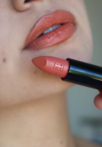 Naakte roze lippenstift dicht bij de lippen gehouden; de tube heeft een zwarte behuizing en een gegraveerd logo; gladde textuur op de lippen met een matte afwerking.
