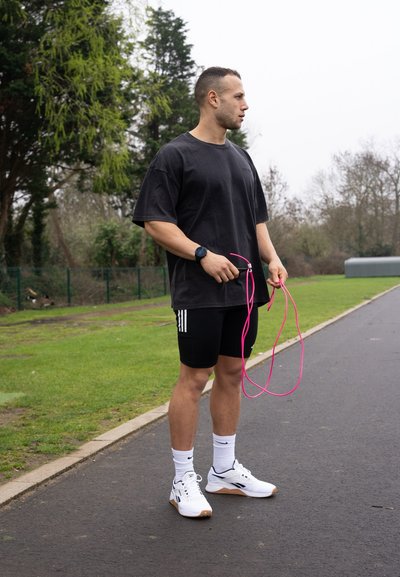 Camiseta negra de algodón, pantalones cortos deportivos negros con rayas blancas, zapatillas blancas, sosteniendo una cuerda de saltar rosa, de pie en un camino pavimentado.