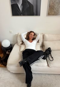 Haut blanc à manches longues et pantalon de survêtement noir avec des rayures blanches. Assise sur un canapé de couleur claire, sac à main noir à proximité, et une lampe ronde blanche.