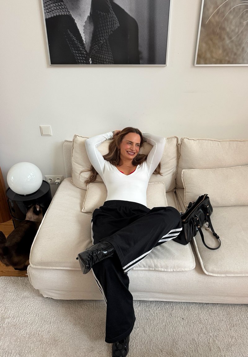 Haut blanc à manches longues et pantalon de survêtement noir avec des rayures blanches. Assise sur un canapé de couleur claire, sac à main noir à proximité, et une lampe ronde blanche.