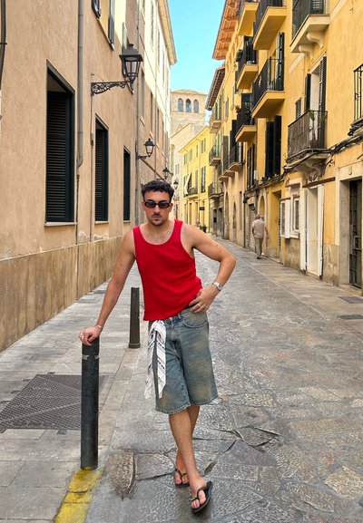 Top rojo de tirantes, pantalones cortos de mezclilla, sandalias grises. De pie en una calle estrecha con edificios amarillos y beige, pavimento de piedra y farolas.