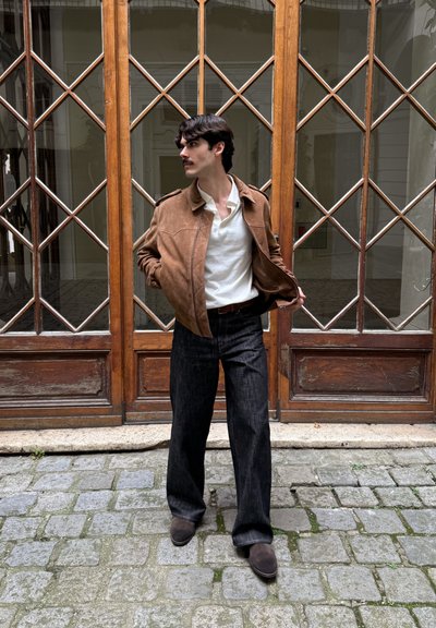 Chaqueta bomber de ante marrón sobre una camisa blanca de cuello, combinada con pantalones de mezclilla anchos oscuros y zapatos marrones, frente a unas puertas dobles de madera.