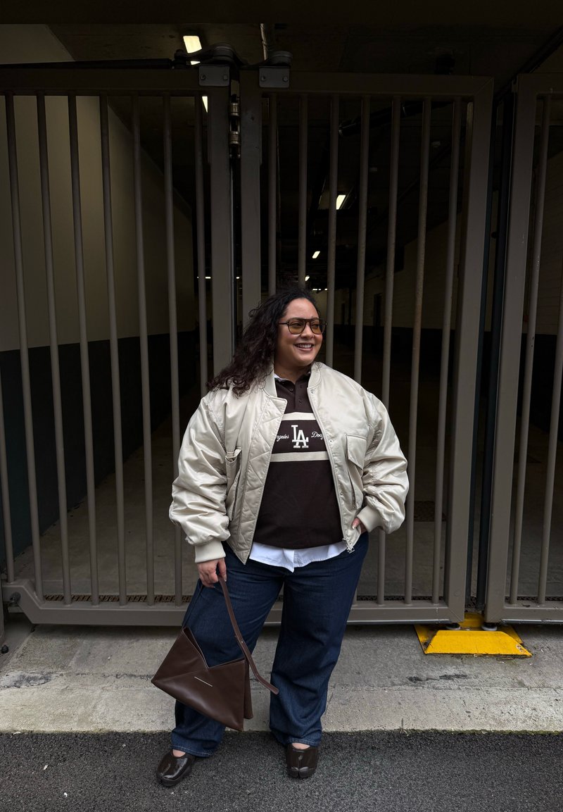 Femme aux cheveux bouclés portant des lunettes, une veste bomber beige, un t-shirt sombre des LA Dodgers, un jean, et tenant un sac à main marron devant une grille métallique.