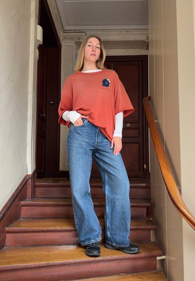Mujer joven de pie en unas escaleras de madera, con jeans vaqueros relajados, camisetas superpuestas en naranja y blanco, con una mano en el bolsillo, en el interior junto a una puerta.