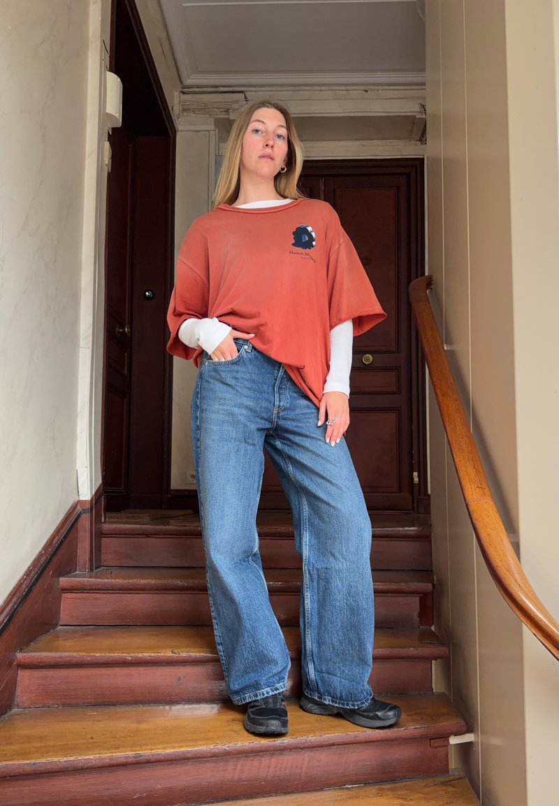 Jonge vrouw die op houten trappen staat, gekleed in relaxte spijkerbroek en laagjes van oranje en witte shirts, met één hand in haar zak, binnen bij een deur.