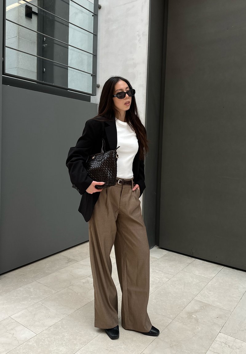 Frau mit Sonnenbrille, schwarzem Blazer, weißem Oberteil und weiten braunen Hosen, die eine schwarze geflochtene Handtasche hält und drinnen neben einer großen grauen Tür steht.